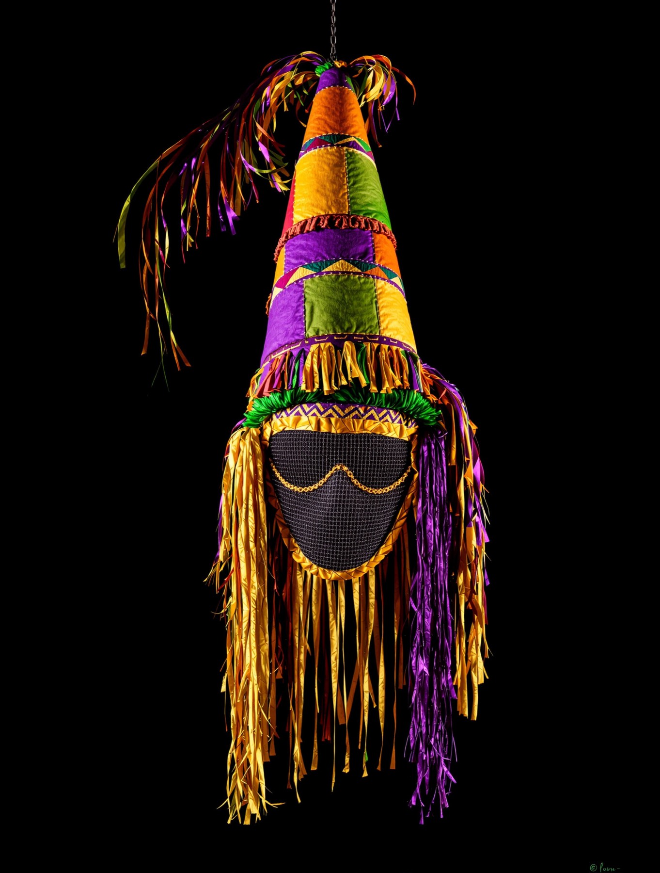 A tall conical fringed Cajun capuchon in bright patchwork fabric with a screen wire mask.